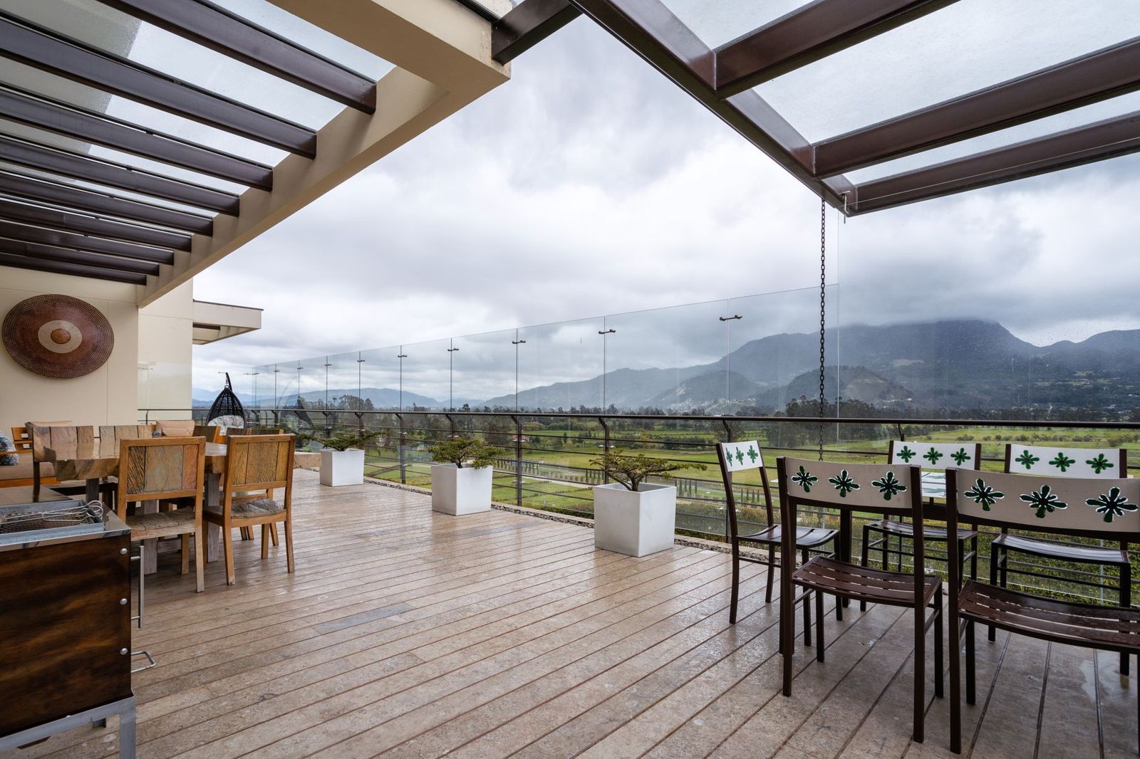 Long covered deck with pale plank decking, glass railing and dining furniture looking out over green hills on a cloudy day