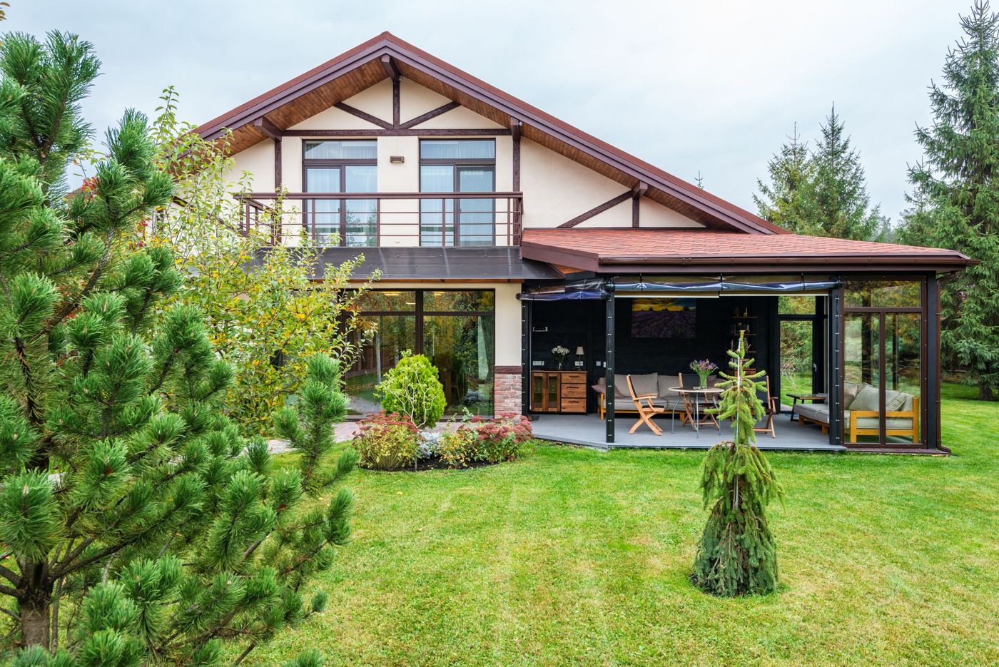 Two-story house with a covered corner patio, outdoor sofa and chairs, and a wide lawn edged by pine trees