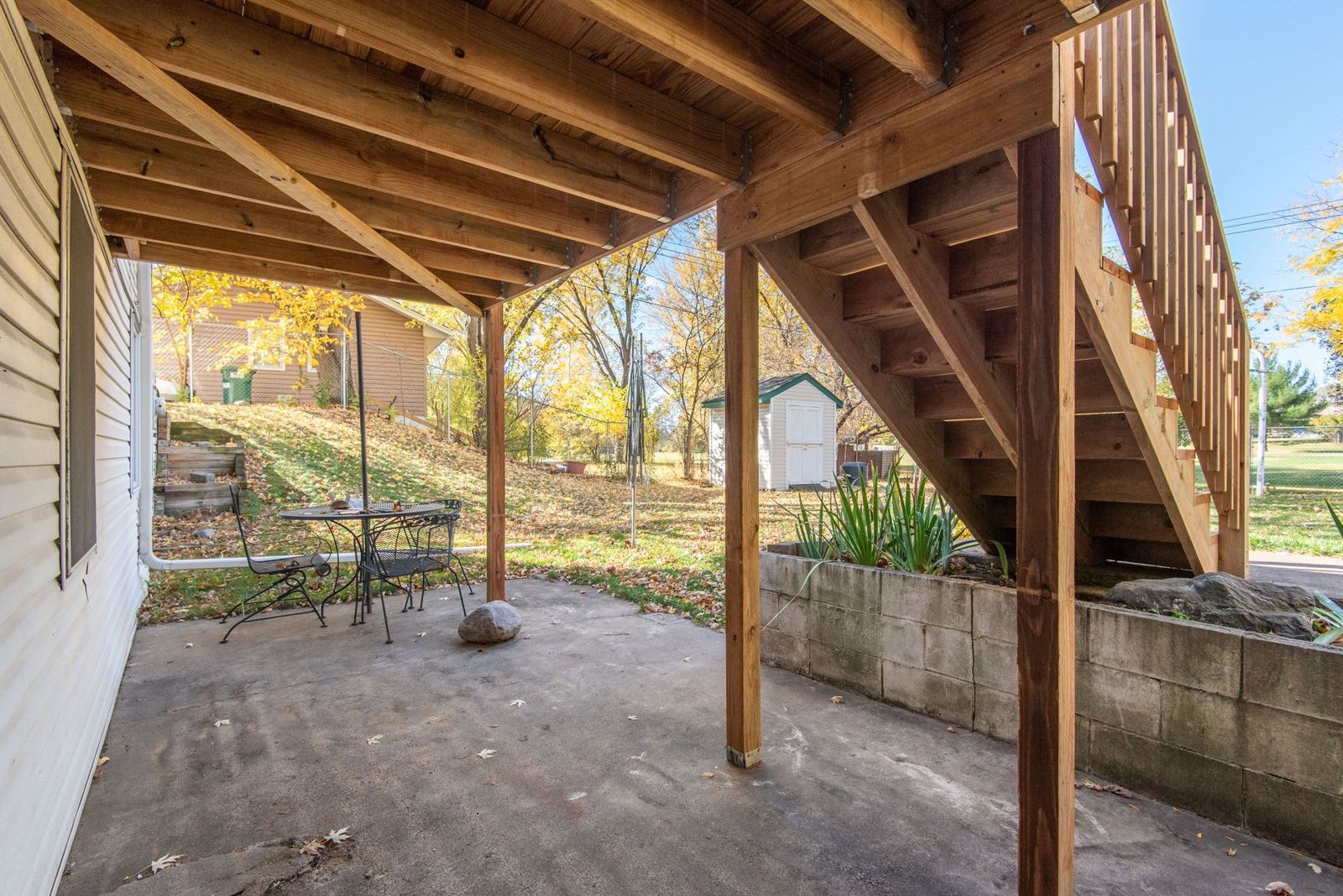 Underside of a raised wooden deck showing joists, beams, support posts and an attached staircase above a concrete patio