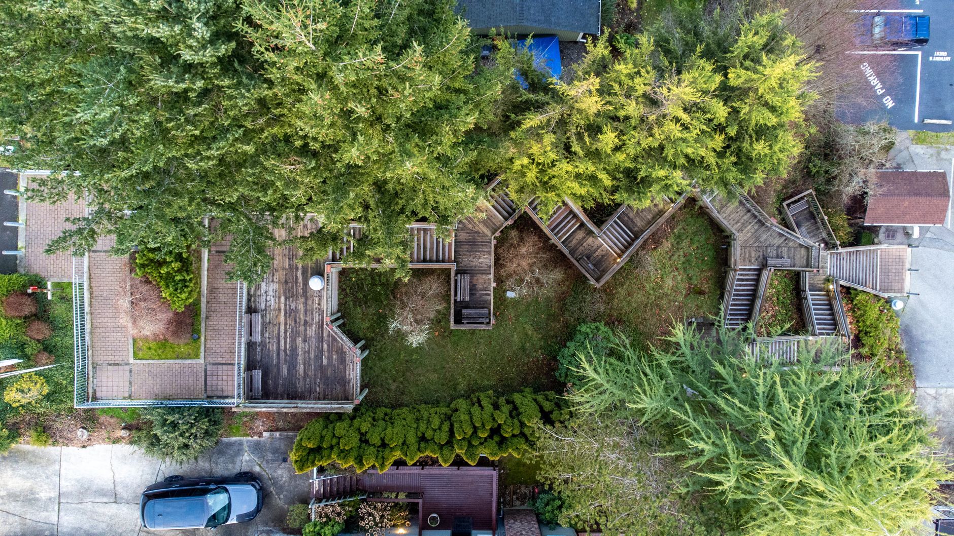 Aerial view of a weathered wooden deck and zigzagging stairs winding through a backyard lawn surrounded by tall evergreens