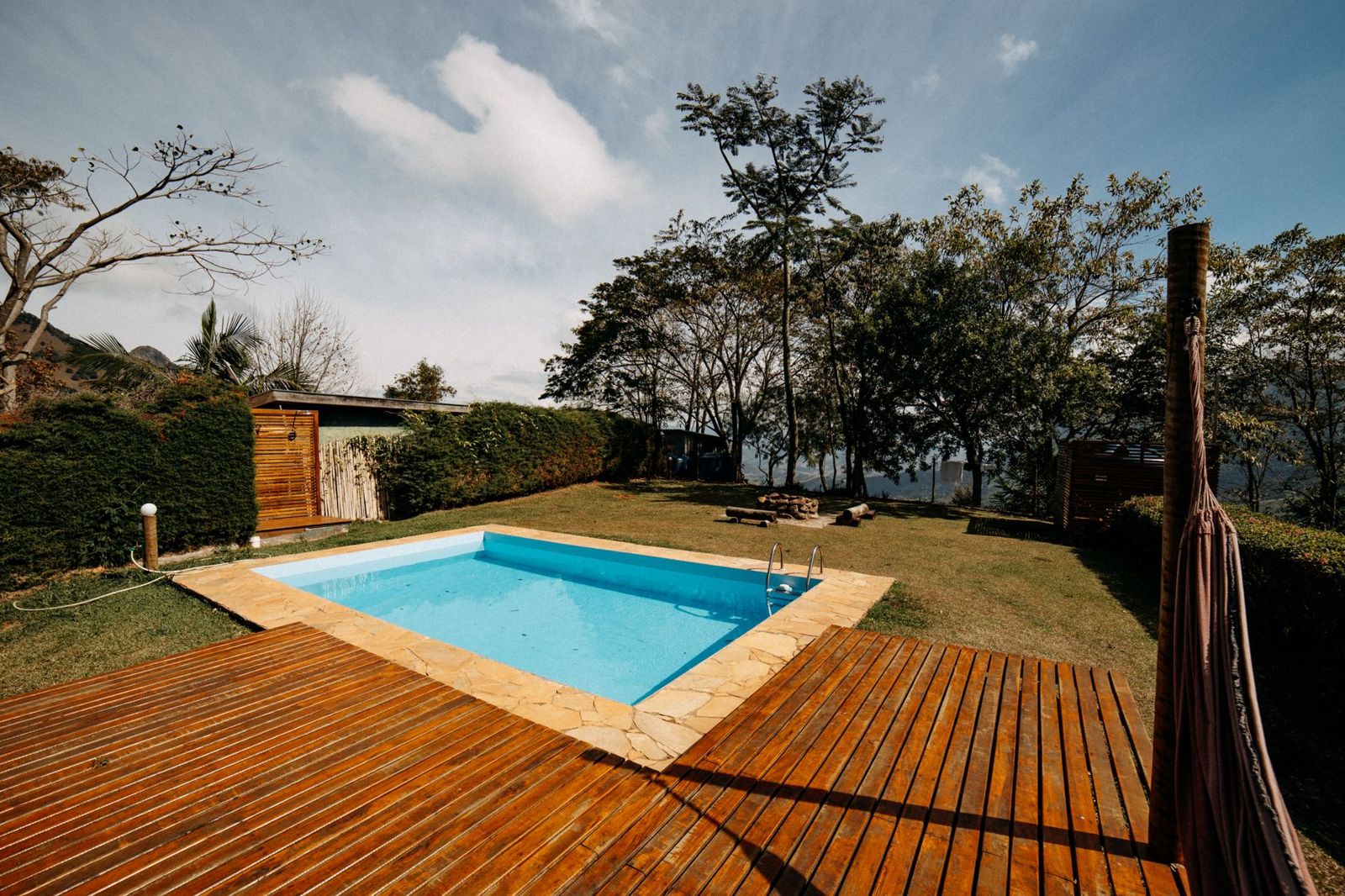 Warm-toned wooden plank deck wrapping a stone-edged swimming pool, with trees and a hammock post