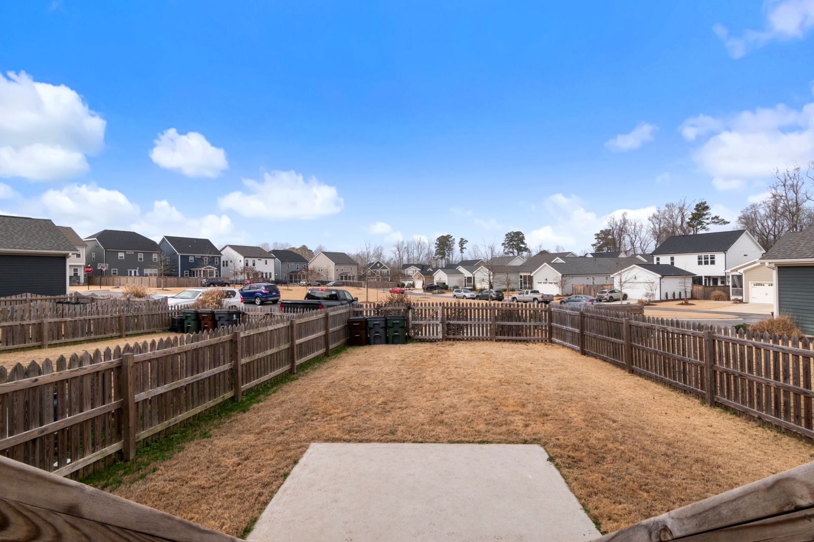 View over a backyard enclosed by weathered wooden picket-style fencing, with a row of suburban houses beyond